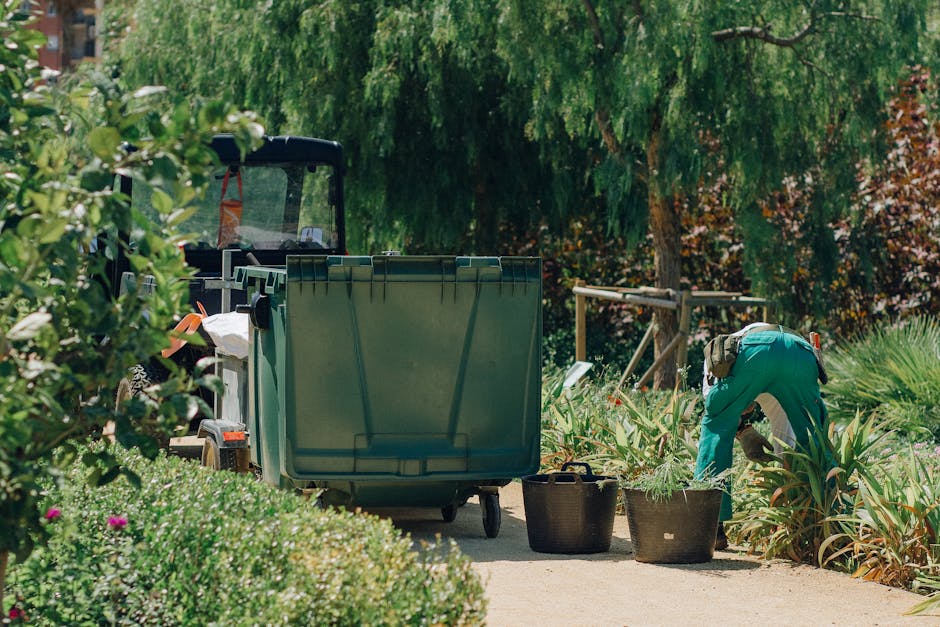A gardener wearing a green uniform is kneeling and trimming plants along a garden pathway. The scene includes a large green waste bin positioned on the paved surface, with additional black baskets nea
