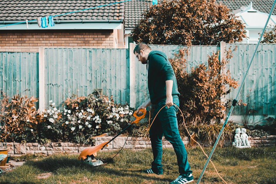A gardener wearing a green uniform is kneeling and trimming plants along a garden pathway. The scene includes a large green waste bin positioned on the paved surface, with additional black baskets nea